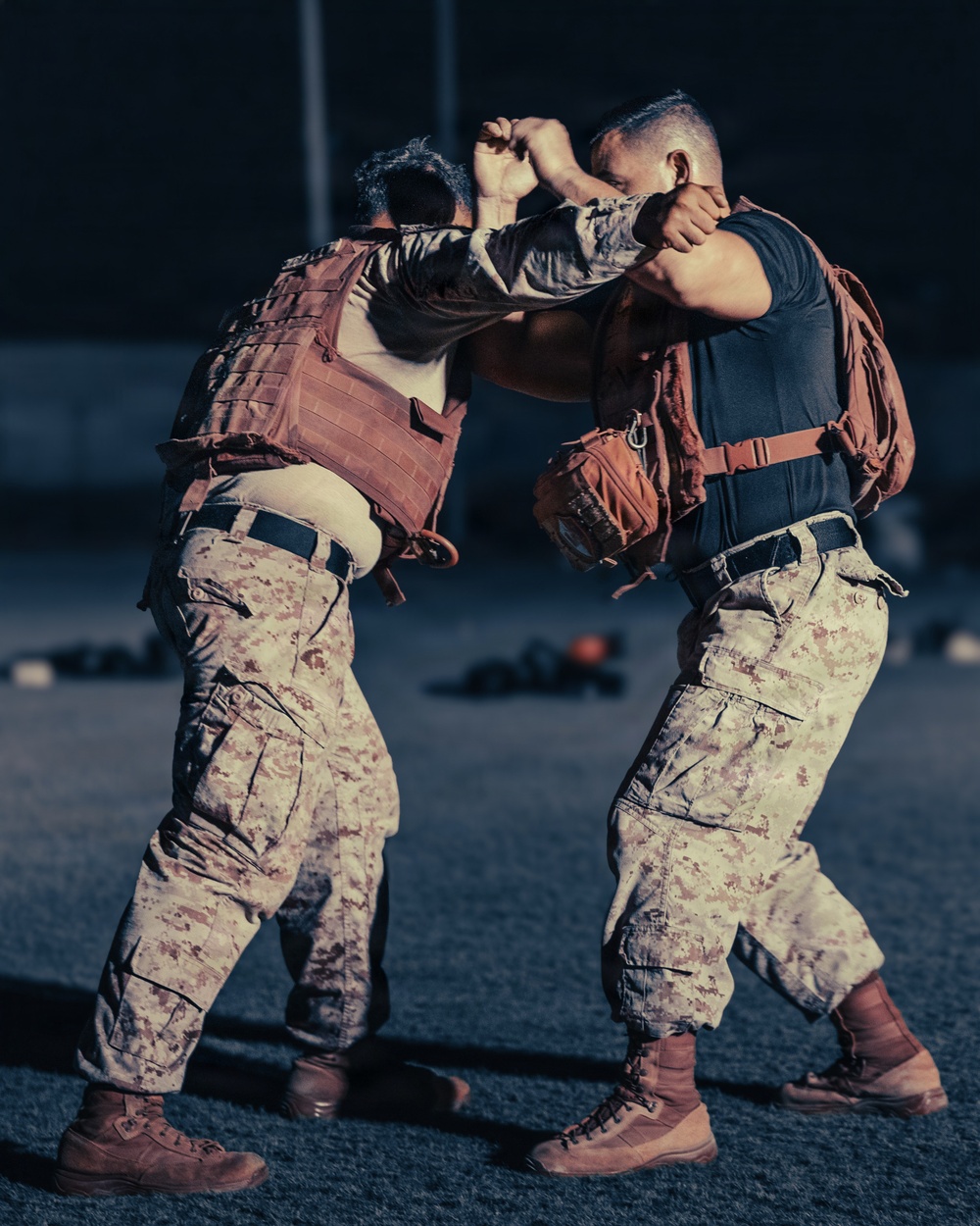 Two individuals in matching desert digital camouflage uniforms are engaged in close-quarters combat training. Both wear tactical gear including plate carriers and backpacks, suggesting a structured military martial arts or combatives drill. The image is captured in a dramatic low-light outdoor setting, likely on a training field.