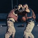 Two individuals in matching desert digital camouflage uniforms are engaged in close-quarters combat training. Both wear tactical gear including plate carriers and backpacks, suggesting a structured military martial arts or combatives drill. The image is captured in a dramatic low-light outdoor setting, likely on a training field.