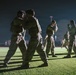Military personnel in camouflage uniforms and tactical vests are conducting hand-to-hand combat training on a grass field at night. Multiple pairs of soldiers are practicing close-quarters fighting techniques simultaneously under artificial lighting. The scene appears to be a structured martial arts or combatives training exercise on what looks like a military installation.