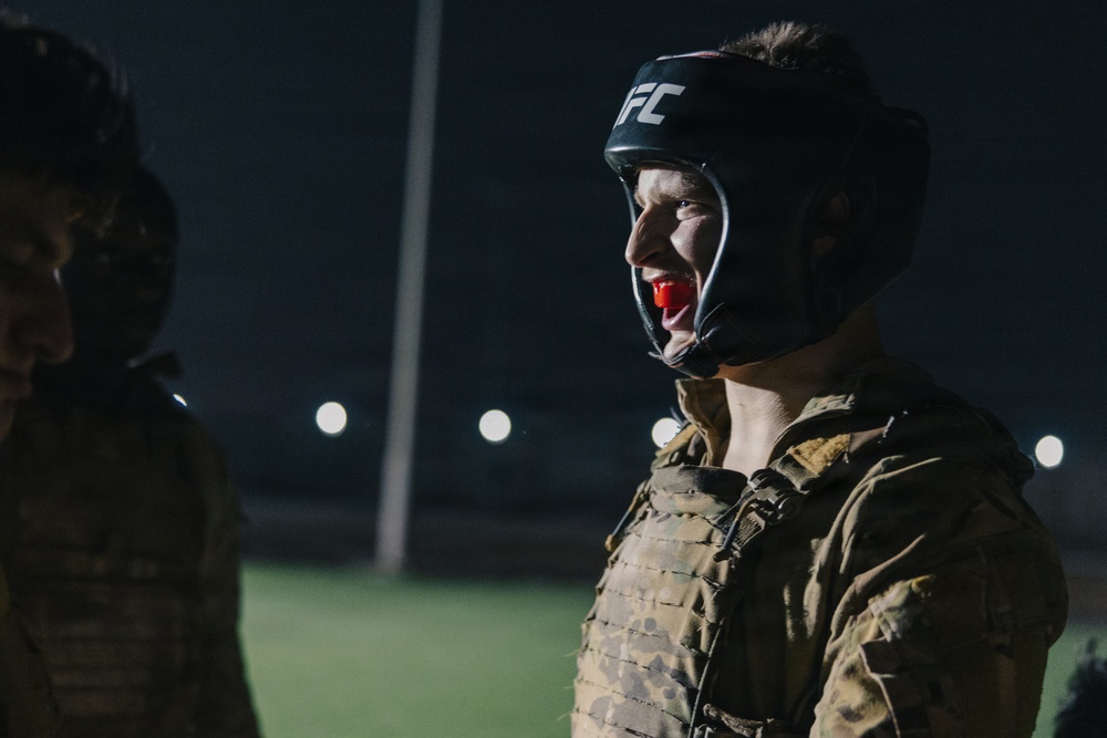 A person wearing military camouflage uniform and a UFC-branded black head guard with a red mouthguard is photographed at night under artificial lighting. The individual appears to be in a combat sports training or sparring context, likely a military combatives exercise. The background shows a lit field or outdoor training area.