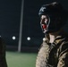 A person wearing military camouflage uniform and a UFC-branded black head guard with a red mouthguard is photographed at night under artificial lighting. The individual appears to be in a combat sports training or sparring context, likely a military combatives exercise. The background shows a lit field or outdoor training area.