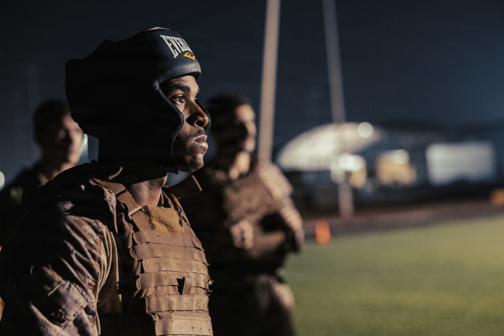 A person wearing an Everlast boxing headgear and a tactical/plate carrier vest stands at attention or in formation during what appears to be a nighttime military or law enforcement training exercise. The subject is in sharp focus in the foreground while additional similarly equipped individuals are visible but blurred in the background. The setting appears to be an outdoor field or training ground with structures visible in the far background.