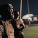 A person wearing an Everlast boxing headgear and a tactical/plate carrier vest stands at attention or in formation during what appears to be a nighttime military or law enforcement training exercise. The subject is in sharp focus in the foreground while additional similarly equipped individuals are visible but blurred in the background. The setting appears to be an outdoor field or training ground with structures visible in the far background.