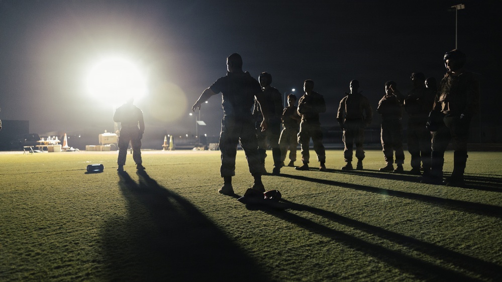 A nighttime military training or briefing session taking place on a grass field, illuminated by a single bright floodlight. Multiple personnel in military/tactical uniforms are gathered in a loose formation, with one individual appearing to address or instruct the group. Long shadows are cast across the field due to the single-point light source.
