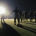A nighttime military training or briefing session taking place on a grass field, illuminated by a single bright floodlight. Multiple personnel in military/tactical uniforms are gathered in a loose formation, with one individual appearing to address or instruct the group. Long shadows are cast across the field due to the single-point light source.