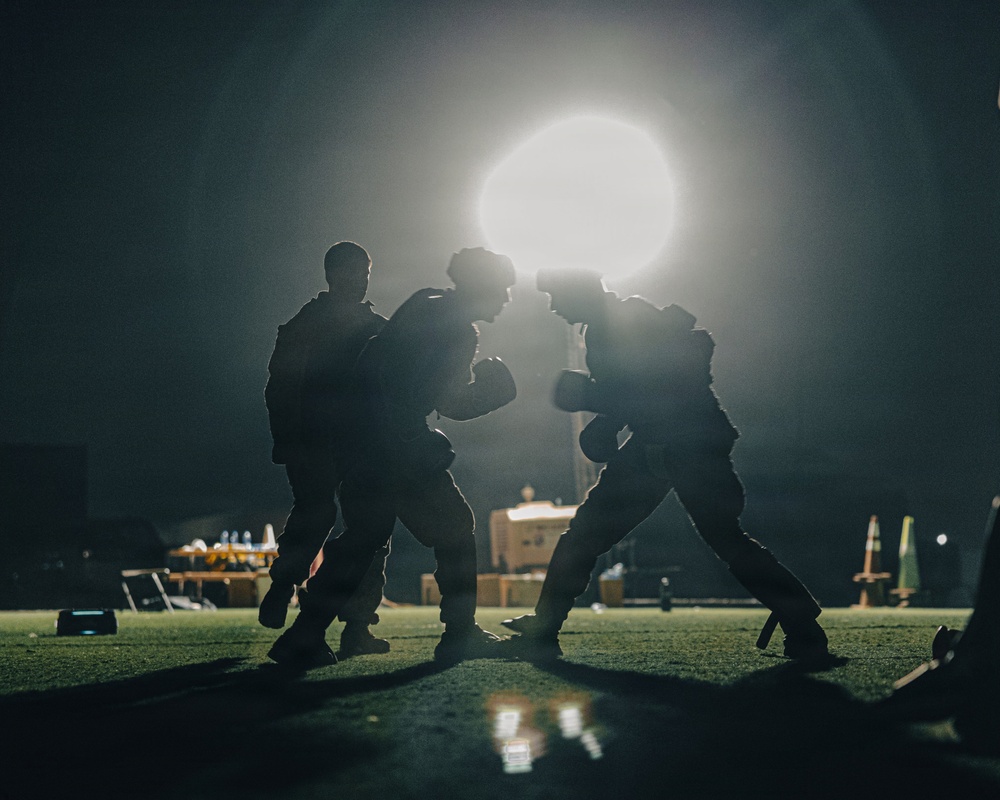 A dramatic nighttime photograph showing silhouetted figures engaged in what appears to be a boxing or combat training session. The scene is backlit by a single powerful light source creating a glowing halo effect. The dark environment and silhouette composition give the image a cinematic, high-contrast aesthetic.