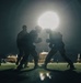 A dramatic nighttime photograph showing silhouetted figures engaged in what appears to be a boxing or combat training session. The scene is backlit by a single powerful light source creating a glowing halo effect. The dark environment and silhouette composition give the image a cinematic, high-contrast aesthetic.