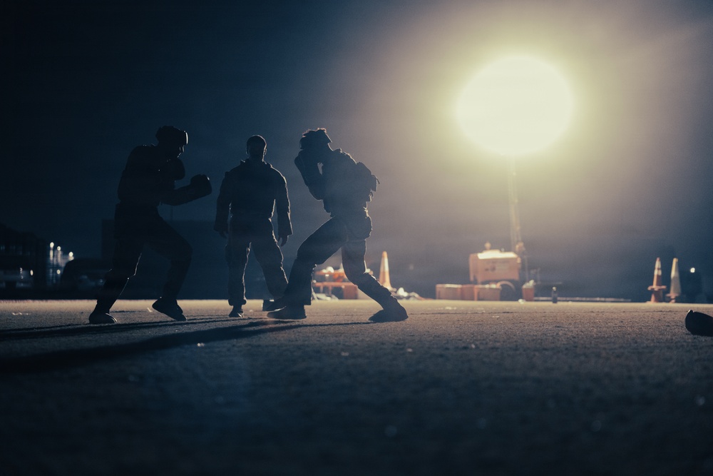 A nighttime scene showing three silhouetted figures engaged in what appears to be a combat training or martial arts sparring session on an open paved surface. The figures are backlit by a large, bright artificial light source creating dramatic silhouette effects. The setting appears to be a military base, airfield, or industrial area based on visible background equipment.