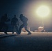 A nighttime scene showing three silhouetted figures engaged in what appears to be a combat training or martial arts sparring session on an open paved surface. The figures are backlit by a large, bright artificial light source creating dramatic silhouette effects. The setting appears to be a military base, airfield, or industrial area based on visible background equipment.
