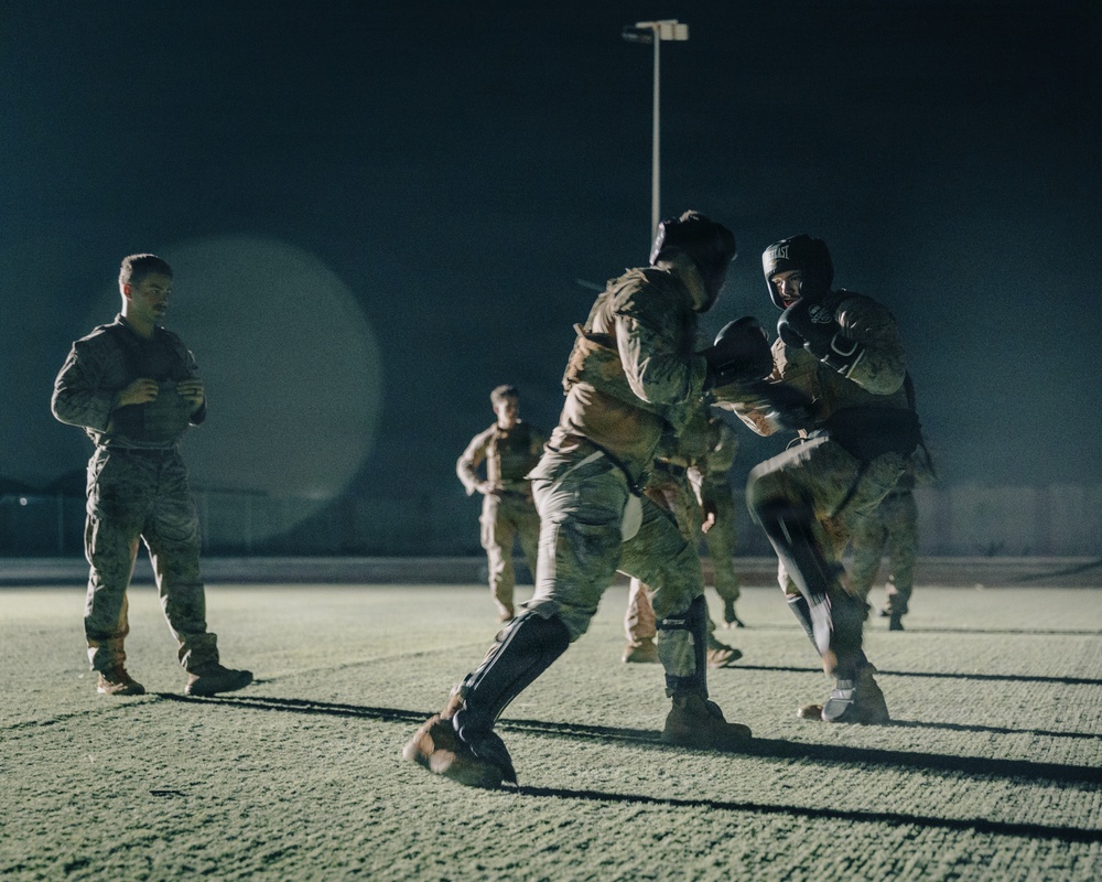 A nighttime military combatives training session taking place on an outdoor paved surface, illuminated by artificial lighting. Two soldiers in the foreground are actively sparring while wearing protective gear including boxing gloves and helmets, while other personnel observe or participate in the background. A supervising soldier stands to the left with arms crossed, monitoring the exercise.