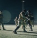 A nighttime military combatives training session taking place on an outdoor paved surface, illuminated by artificial lighting. Two soldiers in the foreground are actively sparring while wearing protective gear including boxing gloves and helmets, while other personnel observe or participate in the background. A supervising soldier stands to the left with arms crossed, monitoring the exercise.