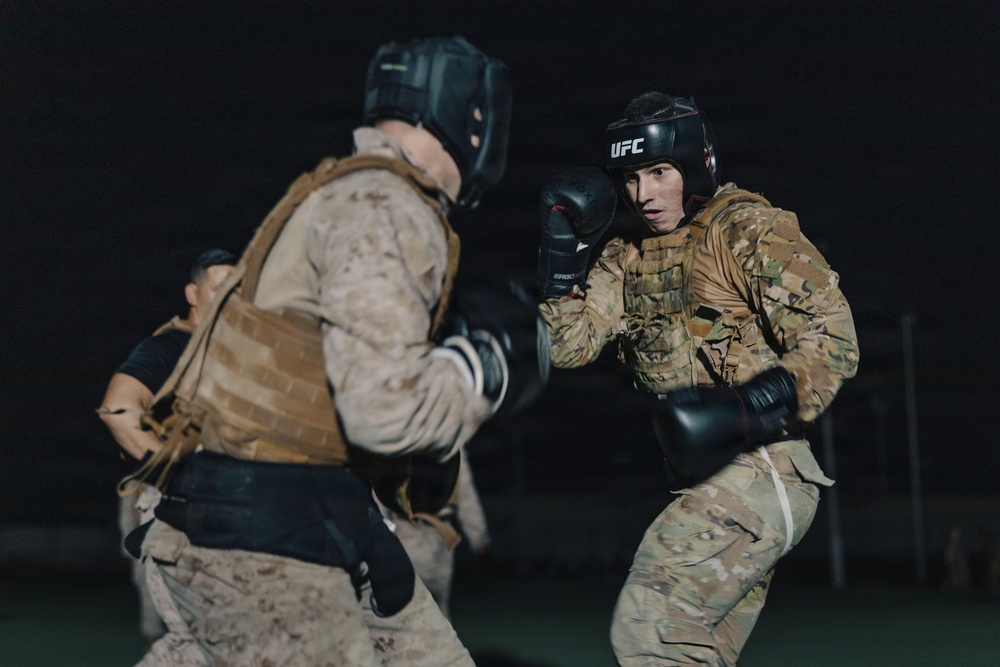 Two military personnel are engaged in a combat training exercise at night, wearing full combat uniforms with tactical vests and boxing protective gear. The individual on the right wears a UFC-branded headguard and black boxing gloves in a fighting stance, facing an opponent in desert camouflage. A third person is visible in the background observing or participating in the drill.