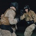 Two military personnel are engaged in a combat training exercise at night, wearing full combat uniforms with tactical vests and boxing protective gear. The individual on the right wears a UFC-branded headguard and black boxing gloves in a fighting stance, facing an opponent in desert camouflage. A third person is visible in the background observing or participating in the drill.