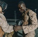 Two individuals in U.S. Marine Corps MARPAT desert camouflage uniforms and tactical vests are preparing for a combat training exercise at night. One person is assisting the other in putting on or adjusting a 12oz boxing glove, while the other wears a protective headgear. The setting appears to be an outdoor training area illuminated by artificial lighting.