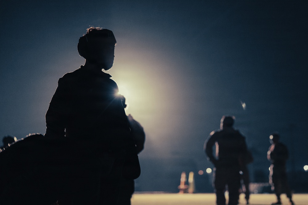 A nighttime scene showing silhouetted figures against a bright artificial light source, creating a dramatic backlit composition. The foreground features a prominently silhouetted individual, while additional figures are visible in the middle and background distance. The atmospheric haze around the light source suggests fog, dust, or humid conditions.