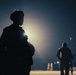 A nighttime scene showing silhouetted figures against a bright artificial light source, creating a dramatic backlit composition. The foreground features a prominently silhouetted individual, while additional figures are visible in the middle and background distance. The atmospheric haze around the light source suggests fog, dust, or humid conditions.