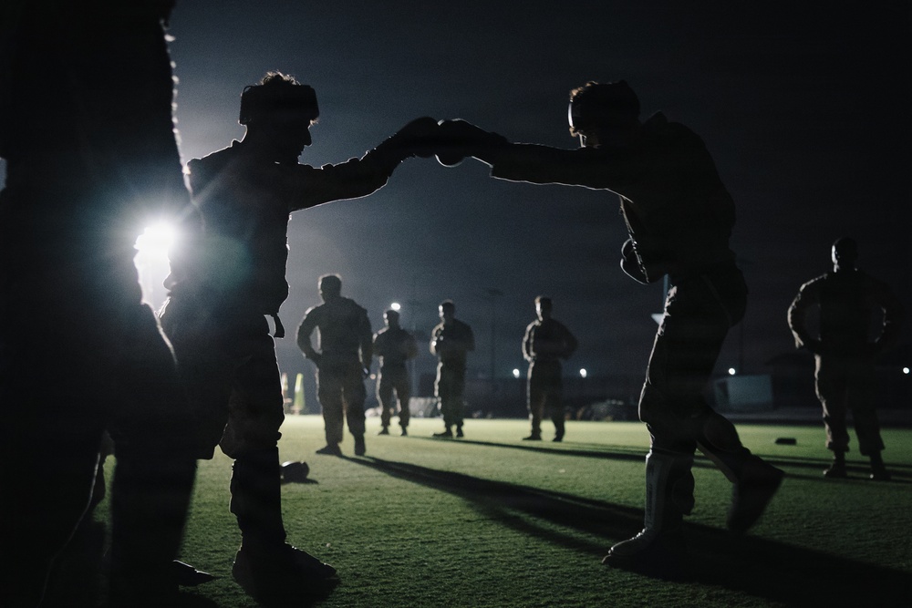 A nighttime outdoor scene on an artificial turf field showing multiple individuals in military-style uniforms engaged in what appears to be combatives or hand-to-hand combat training. Two figures in the foreground are prominently silhouetted, appearing to spar or practice defensive techniques while wearing helmets and protective gear. Several other uniformed personnel are visible in the background under artificial lighting.