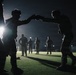 A nighttime outdoor scene on an artificial turf field showing multiple individuals in military-style uniforms engaged in what appears to be combatives or hand-to-hand combat training. Two figures in the foreground are prominently silhouetted, appearing to spar or practice defensive techniques while wearing helmets and protective gear. Several other uniformed personnel are visible in the background under artificial lighting.