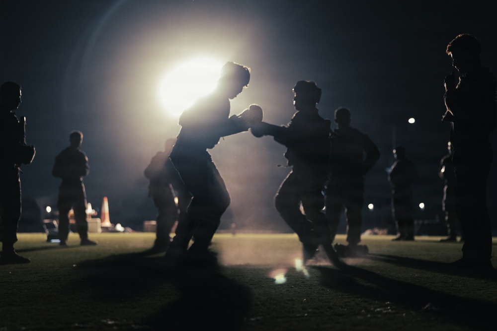 A nighttime outdoor scene showing two individuals engaged in a boxing or combat training sparring session, backlit by a bright artificial light source. Several other figures stand around the perimeter observing, suggesting a supervised training exercise. The dramatic backlighting creates strong silhouettes against the dark environment.