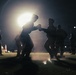 A nighttime outdoor scene showing two individuals engaged in a boxing or combat training sparring session, backlit by a bright artificial light source. Several other figures stand around the perimeter observing, suggesting a supervised training exercise. The dramatic backlighting creates strong silhouettes against the dark environment.