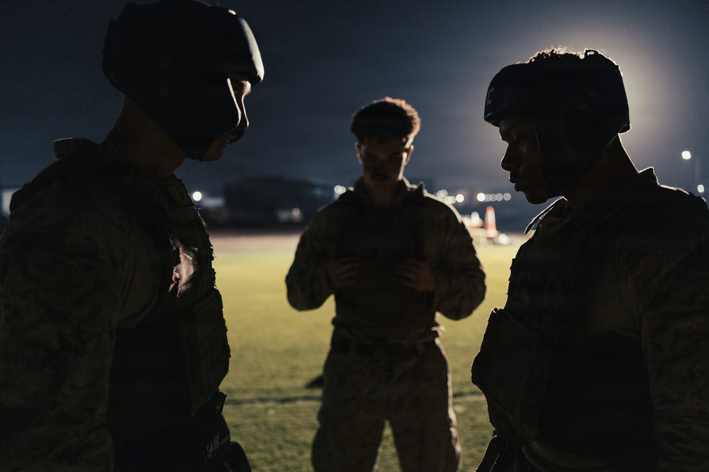 Three individuals in military camouflage uniforms are gathered in a huddle during nighttime operations. Two of the figures are wearing tactical helmets while the central figure appears to be reviewing something, possibly a device or document. The scene is set on what appears to be a military installation or airfield, illuminated by distant artificial lighting.