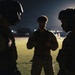 Three individuals in military camouflage uniforms are gathered in a huddle during nighttime operations. Two of the figures are wearing tactical helmets while the central figure appears to be reviewing something, possibly a device or document. The scene is set on what appears to be a military installation or airfield, illuminated by distant artificial lighting.