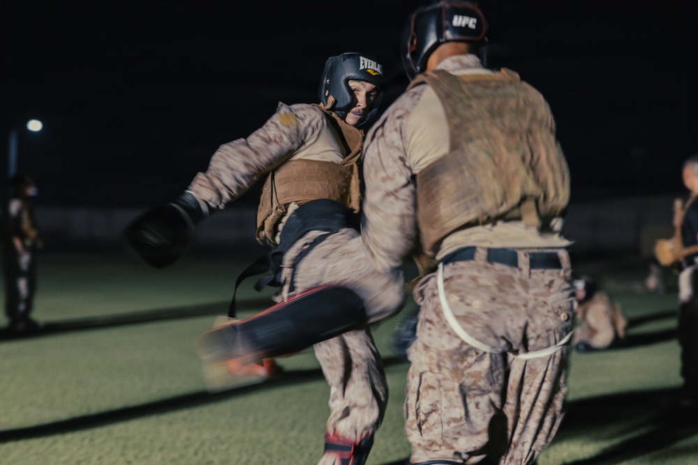 Two individuals in military camouflage uniforms and protective gear are engaged in a combat training exercise at night on what appears to be an outdoor field. The person on the left is executing a kick while wearing an Everlast boxing headguard, while the opponent wears a UFC-branded headguard. Additional participants are visible in the blurred background, suggesting a group training session.