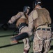 Two individuals in military camouflage uniforms and protective gear are engaged in a combat training exercise at night on what appears to be an outdoor field. The person on the left is executing a kick while wearing an Everlast boxing headguard, while the opponent wears a UFC-branded headguard. Additional participants are visible in the blurred background, suggesting a group training session.