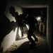 A low-light image showing military personnel moving through a narrow, dark corridor or hallway with sandy floors. At least two figures in tactical gear are moving in a crouched, dynamic posture, while a third individual holding a flashlight stands in a doorway. The scene appears to depict a military training exercise or operational building clearance.