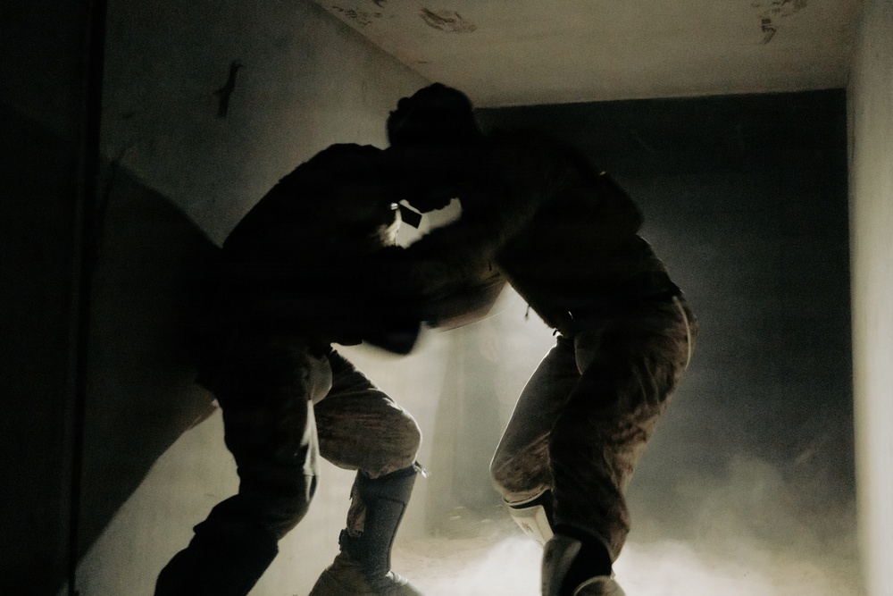 Two individuals wearing military or tactical gear are visible in a dark, confined interior space filled with smoke or dust. The figures are heavily backlit, creating strong silhouettes against a bright light source emanating from below or behind them. The environment appears to be a damaged or deteriorated building interior.
