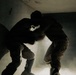Two individuals wearing military or tactical gear are visible in a dark, confined interior space filled with smoke or dust. The figures are heavily backlit, creating strong silhouettes against a bright light source emanating from below or behind them. The environment appears to be a damaged or deteriorated building interior.