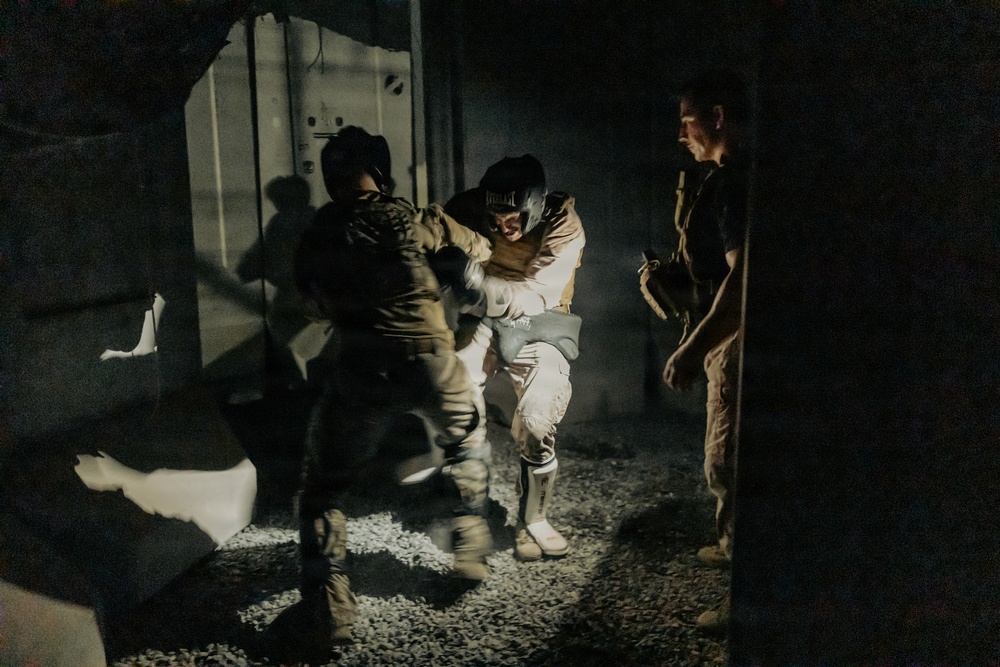 A low-light nighttime scene showing individuals in military or tactical gear in what appears to be a confined space or building interior. Two individuals appear to be engaged in close physical contact, possibly a training exercise, apprehension, or assistance scenario. A third individual stands nearby observing.