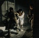A low-light nighttime scene showing individuals in military or tactical gear in what appears to be a confined space or building interior. Two individuals appear to be engaged in close physical contact, possibly a training exercise, apprehension, or assistance scenario. A third individual stands nearby observing.