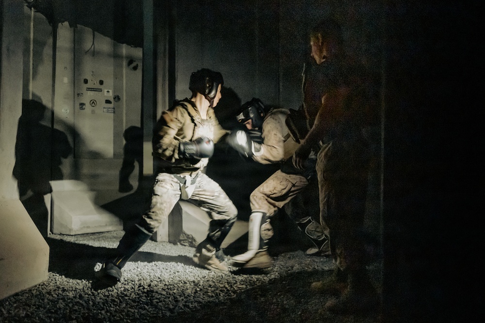 Two individuals in military-style camouflage uniforms and protective headgear are engaged in a close-quarters combat or martial arts sparring session in a low-light outdoor or semi-enclosed environment. A third person stands nearby observing the activity. The scene appears to be a training exercise based on the protective equipment worn.