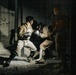 Two individuals in military-style camouflage uniforms and protective headgear are engaged in a close-quarters combat or martial arts sparring session in a low-light outdoor or semi-enclosed environment. A third person stands nearby observing the activity. The scene appears to be a training exercise based on the protective equipment worn.