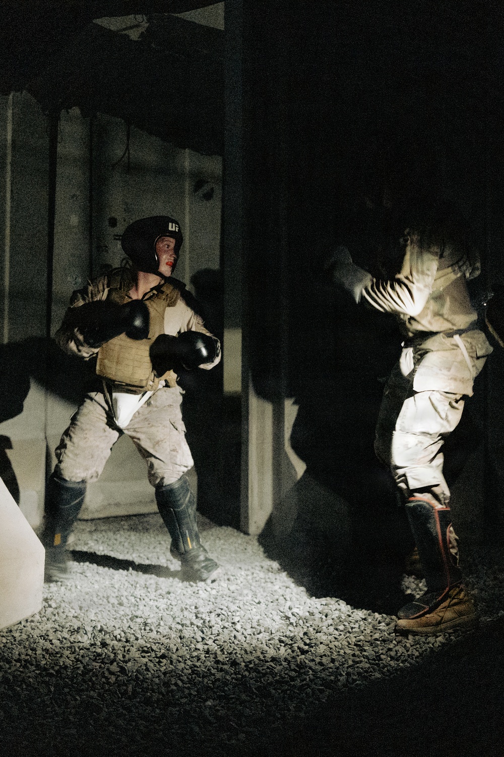 Two individuals are engaged in a combat training exercise in a dark, confined space illuminated by a spotlight. The left figure wears military-style tactical gear including a plate carrier vest, boxing gloves, a UFC-branded head guard, and combat boots, suggesting a military hand-to-hand combat drill. The right figure appears to be a padded training partner or opponent in protective equipment.