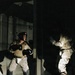 Two individuals are engaged in a combat training exercise in a dark, confined space illuminated by a spotlight. The left figure wears military-style tactical gear including a plate carrier vest, boxing gloves, a UFC-branded head guard, and combat boots, suggesting a military hand-to-hand combat drill. The right figure appears to be a padded training partner or opponent in protective equipment.