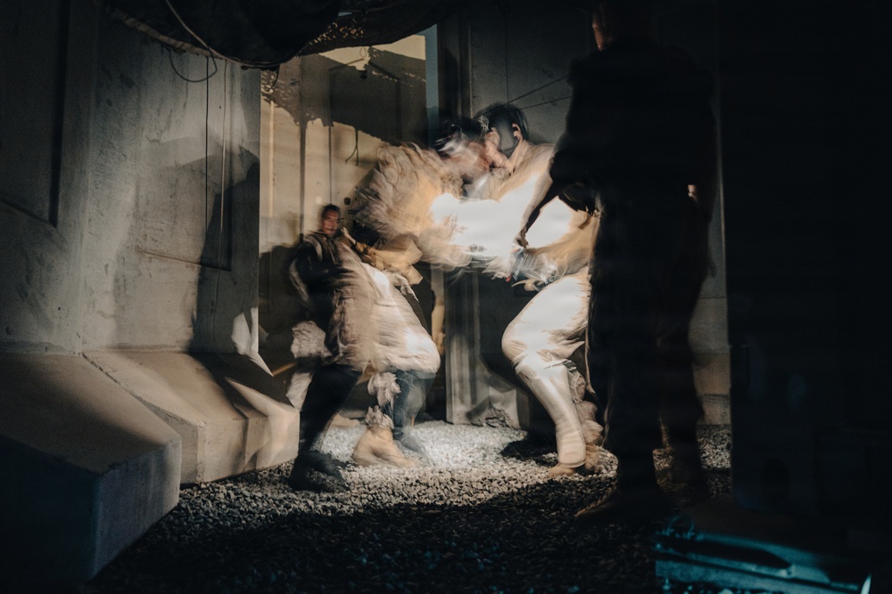 A long-exposure or motion-blurred photograph captures figures in white/light-colored costumes or protective suits engaged in dynamic movement within an industrial or brutalist concrete space. The motion blur creates ghostly, layered impressions of the figures, suggesting performance art, dance, or a staged theatrical event. The ground is covered in gravel or crushed stone, and the setting appears to be a raw, unfinished architectural environment.