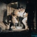 A long-exposure or motion-blurred photograph captures figures in white/light-colored costumes or protective suits engaged in dynamic movement within an industrial or brutalist concrete space. The motion blur creates ghostly, layered impressions of the figures, suggesting performance art, dance, or a staged theatrical event. The ground is covered in gravel or crushed stone, and the setting appears to be a raw, unfinished architectural environment.