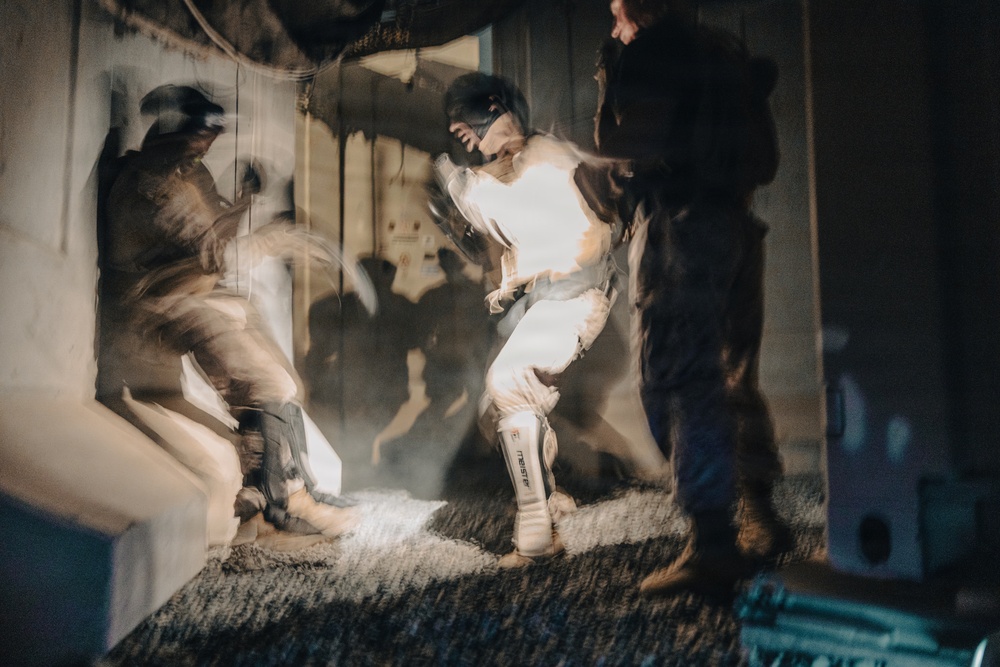 A long-exposure or multiple-exposure photograph capturing what appears to be a martial arts or combat sports training session in an indoor space. The motion blur creates ghosting effects showing the movement of at least two fighters, with one wearing white gear including visible shin guards. The artistic technique results in layered, translucent figures suggesting dynamic striking or sparring activity.