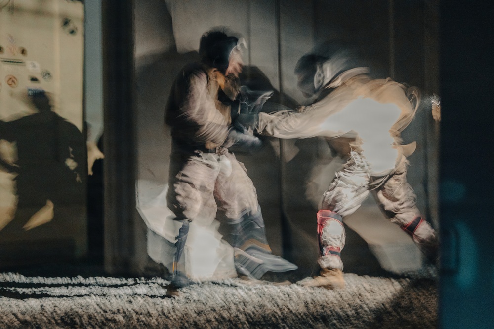 A long-exposure or motion-blur photograph captures two individuals engaged in a martial arts sparring session, likely karate or taekwondo, indoors on a mat. The intentional camera movement or slow shutter speed creates ghosting effects that show multiple positions of the fighters simultaneously. Shadows of additional figures are visible on the wall to the left, suggesting other practitioners or spectators are present.