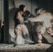 A long-exposure or motion-blur photograph captures two individuals engaged in a martial arts sparring session, likely karate or taekwondo, indoors on a mat. The intentional camera movement or slow shutter speed creates ghosting effects that show multiple positions of the fighters simultaneously. Shadows of additional figures are visible on the wall to the left, suggesting other practitioners or spectators are present.