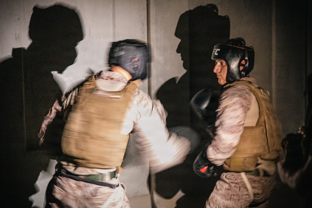 Two individuals in military/tactical gear are engaged in a boxing or combatives sparring session. Both wear protective headgear and boxing gloves along with tactical vests and camouflage uniforms. The motion blur on the left figure suggests an active punch or strike is being thrown toward the right figure.