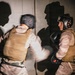 Two individuals in military/tactical gear are engaged in a boxing or combatives sparring session. Both wear protective headgear and boxing gloves along with tactical vests and camouflage uniforms. The motion blur on the left figure suggests an active punch or strike is being thrown toward the right figure.