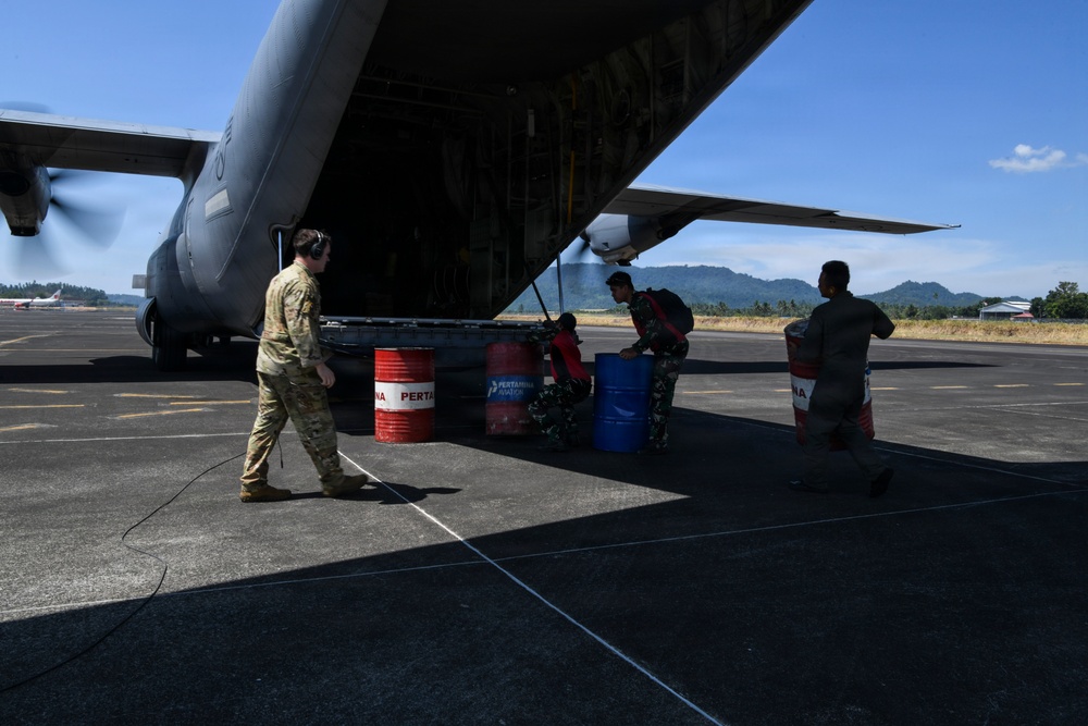 Military personnel are loading or unloading fuel drums from the rear cargo ramp of a large military transport aircraft, likely a C-130 Hercules, on an airfield tarmac. One individual in US military multicam uniform works alongside what appear to be Indonesian military personnel in camouflage uniforms. The barrels are branded with 'Pertamina Aviation,' an Indonesian state-owned fuel company, suggesting this operation is taking place in Indonesia.