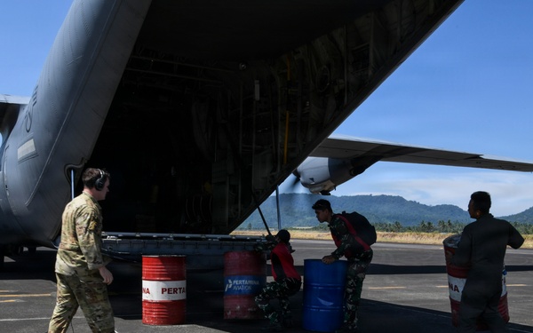 Military personnel are loading or unloading fuel drums from the rear cargo ramp of a large military transport aircraft, likely a C-130 Hercules, on an airfield tarmac. One individual in US military multicam uniform works alongside what appear to be Indonesian military personnel in camouflage uniforms. The barrels are branded with 'Pertamina Aviation,' an Indonesian state-owned fuel company, suggesting this operation is taking place in Indonesia.