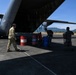 Military personnel are loading or unloading fuel drums from the rear cargo ramp of a large military transport aircraft, likely a C-130 Hercules, on an airfield tarmac. One individual in US military multicam uniform works alongside what appear to be Indonesian military personnel in camouflage uniforms. The barrels are branded with 'Pertamina Aviation,' an Indonesian state-owned fuel company, suggesting this operation is taking place in Indonesia.