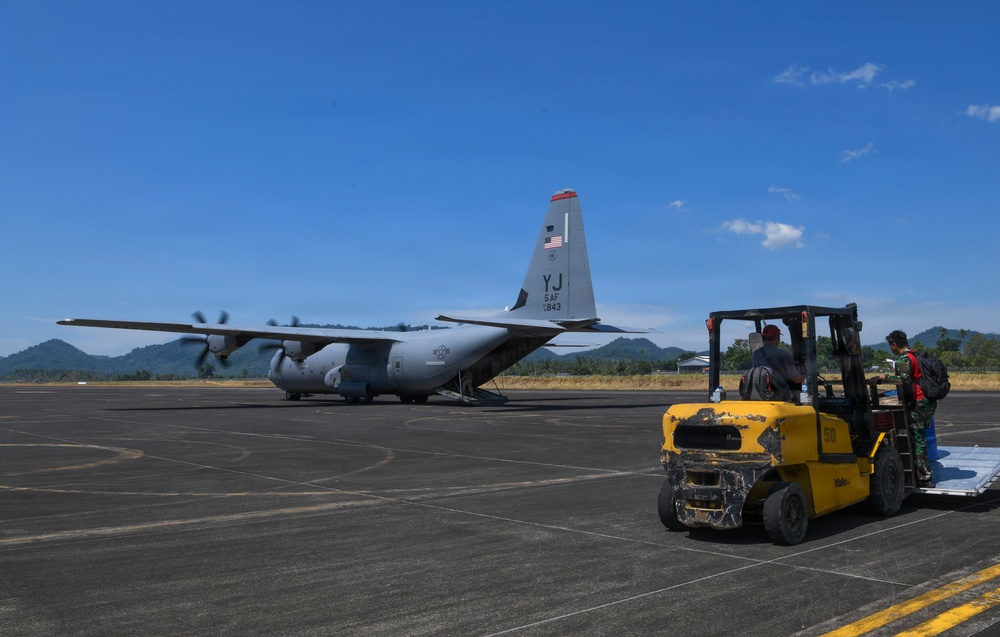 A U.S. Air Force C-130 Hercules transport aircraft, marked with tail code 'YJ' indicating 5th Air Force assignment, is parked on a tarmac with its rear ramp open. Ground personnel are operating a yellow forklift, apparently loading or unloading cargo from the aircraft. The setting appears to be a regional airfield in a tropical or subtropical location, evidenced by the surrounding green hills and vegetation.