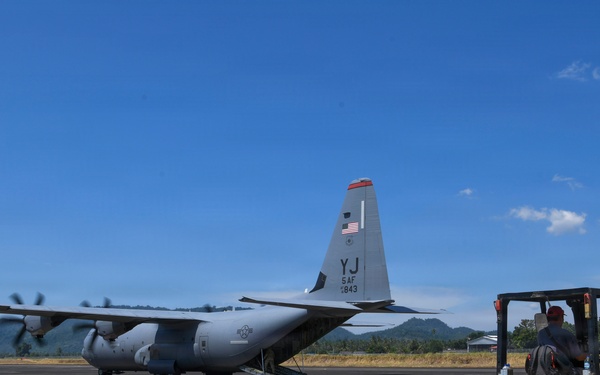 A U.S. Air Force C-130 Hercules transport aircraft, marked with tail code 'YJ' indicating 5th Air Force assignment, is parked on a tarmac with its rear ramp open. Ground personnel are operating a yellow forklift, apparently loading or unloading cargo from the aircraft. The setting appears to be a regional airfield in a tropical or subtropical location, evidenced by the surrounding green hills and vegetation.