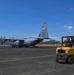 A U.S. Air Force C-130 Hercules transport aircraft, marked with tail code 'YJ' indicating 5th Air Force assignment, is parked on a tarmac with its rear ramp open. Ground personnel are operating a yellow forklift, apparently loading or unloading cargo from the aircraft. The setting appears to be a regional airfield in a tropical or subtropical location, evidenced by the surrounding green hills and vegetation.